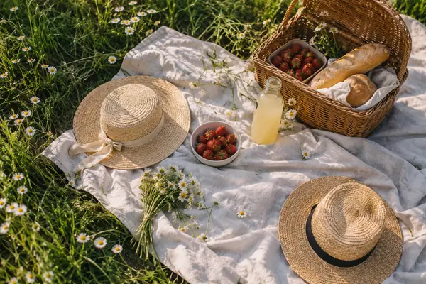 Picknickdecke auf einer Sommerwiese von oben, mit Korb, Erdbeeren, Brot und zwei Strohhüten